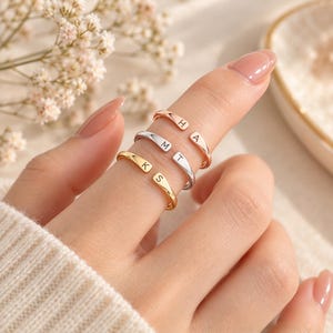 Könnte beinhalten: Drei offene Ringe in Roségold-, Silber- und Goldtönen. Jeder Ring hat zwei flache, abgerundete Enden, die mit einzelnen Buchstaben graviert sind. Der roségoldene Ring zeigt H und A, der silberne Ring M und T und der goldene Ring K und S.