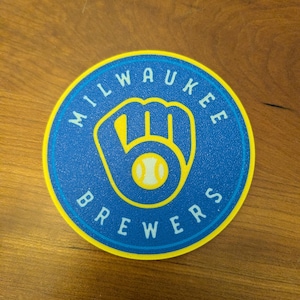 May include: A circular sticker featuring the Milwaukee Brewers logo in yellow and white on a blue background. The text MILWAUKEE BREWERS is printed in white capital letters along the top and bottom edges of the blue circle, bordered by a yellow ring.