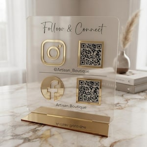 Peut inclure: Un panneau en acrylique transparent avec une base dorée affichant le texte Follow & Connect, des icônes Instagram et Facebook, deux codes QR, le nom @Artisan_Boutique et le texte Artisan Boutique. Ce panneau est destiné aux réseaux sociaux.