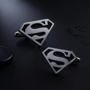 May include: A pair of silver-toned metal cufflinks, each shaped like a diamond with a black-filled Superman S-shield logo. The cufflinks feature a polished metallic finish and a classic T-bar fastening mechanism on the back for secure attachment.