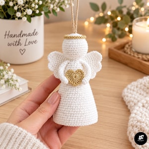 Puede incluir: Un adorno de ángel tejido a ganchillo en color blanco con un halo dorado y un pequeño corazón dorado sostenido entre sus brazos. La figura tiene un cuerpo cónico, cabeza redondeada y alas festoneadas, colgada de un cordel de yute.