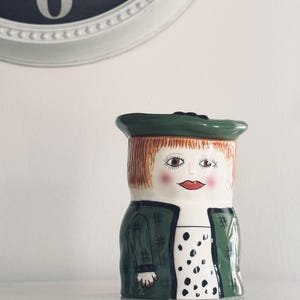 May include: A ceramic jar shaped like a person wearing a green hat, a green jacket with a dark grid pattern, and a white shirt with black polka dots. The figure has light brown hair, brown eyes, rosy cheeks, and red lips, with a glossy finish.
