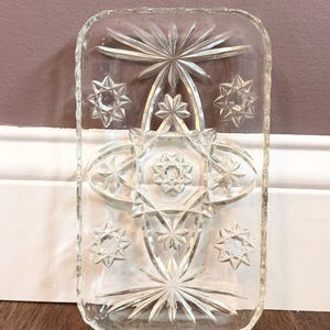 May include: A rectangular clear glass tray featuring an intricate pressed pattern of multiple starbursts and geometric shapes. The edges of the tray have a scalloped, textured finish, and the center displays a prominent central star motif.