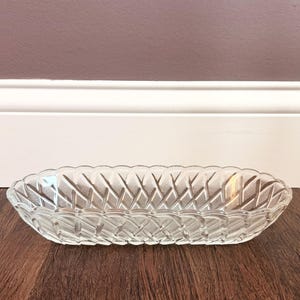 Puede incluir: Un plato ovalado de vidrio transparente que presenta un patrón texturizado de tejido de cesta en su superficie exterior. El plato tiene un borde festoneado decorativo y una base poco profunda y alargada, hecha de vidrio prensado.