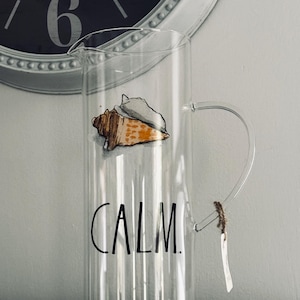 May include: A clear glass pitcher featuring a painted orange and white seashell illustration near the top and the word CALM printed in black capital letters. The pitcher has a curved glass handle with a small brown twine tag attached to it.