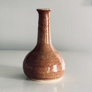 May include: A small, bulbous ceramic bud vase with a glossy, speckled brown glaze. The vase features a narrow, elongated neck that flares slightly at the top opening, with a rounded, wide base that tapers upward into the slender neck structure.
