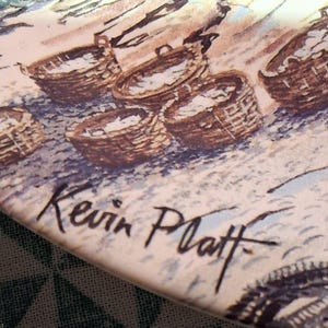 Peut inclure: Une assiette en céramique présentant une illustration détaillée de plusieurs paniers tressés remplis d'objets. L'œuvre est réalisée dans des tons de brun, de beige et de crème. La signature Kevin Platt est visible en encre noire en bas à gauche.