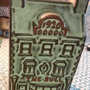 Puede incluir: Una botella de cerámica en forma de casa de color verde claro con detalles en marrón oscuro. El frente presenta un relieve de una fachada con el año 1920, un emblema circular central y el texto THE BULL grabado en letras de color marrón oscuro.
