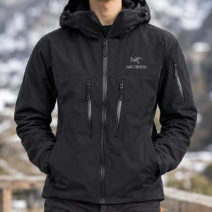 Puede incluir: Una chaqueta negra con capucha que presenta una cremallera frontal completa, dos bolsillos verticales con cremallera en el pecho y un bolsillo con cremallera en la manga izquierda. El logo y nombre Arc'teryx están impresos en blanco.
