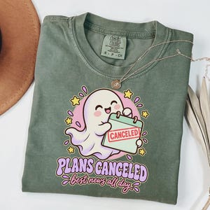 May include: An olive green t-shirt featuring a graphic of a white ghost holding a calendar marked with a red CANCELED stamp. Below the ghost, the text reads PLANS CANCELED best news all day in purple and pink lettering with yellow stars.