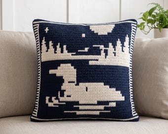 Può includere: Un cuscino quadrato all'uncinetto con uno sfondo blu navy scuro e un motivo grafico color crema. Il disegno raffigura una strolaga che nuota sull'acqua, con la sagoma di un bosco, una luna e stelle sullo sfondo, incorniciato da un bordo a righe.