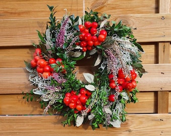 May include: A circular decorative wreath constructed from deep green boxwood foliage, interspersed with clusters of bright red berries, sprigs of purple heather, and pale silvery-green leaves, all arranged in a dense, textured, and natural composition.