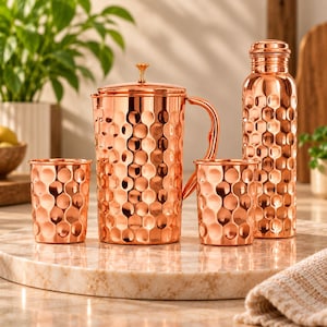 Könnte beinhalten: Ein Set aus poliertem Kupfergeschirr mit gehämmertem Muster. Die Kollektion umfasst einen hohen Krug mit Deckel und Griff, zwei passende Becher sowie eine schlanke Wasserflasche mit Schraubverschluss, die alle eine glänzende Oberfläche aufweisen.