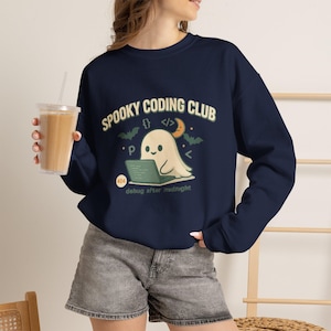 Op de afbeelding: Een marineblauw sweatshirt met lange mouwen en een afbeelding van een geest die een laptop gebruikt. De tekst luidt "SPOOKY CODING CLUB" en "debug after midnight" met een "404"-badge, een halve maan, vleermuizen en diverse codesymbolen.