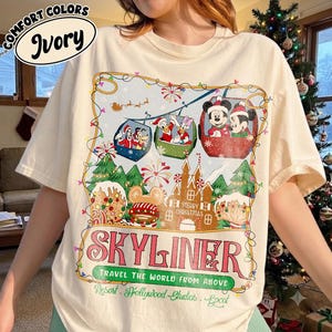 May include: An ivory t-shirt featuring a graphic of Disney characters in holiday-themed skyliner gondolas above a gingerbread castle. The text reads COMFORT COLORS IVORY, SKYLINER, TRAVEL THE WORLD FROM ABOVE, MERRY CHRISTMAS, and Resort Hollywood Studios Epcot.