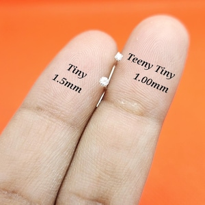 May include: Two small silver-toned stud earrings with clear stones. One earring is labeled Tiny 1.5mm and the other is labeled Teeny Tiny 1.00mm. The earrings are metallic with a prong setting, showcasing a clear, faceted stone on each piece.