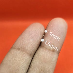 May include: Two small silver stud earrings with clear stones, one labeled 1mm and the other labeled 1.5mm. The earrings are positioned to show their relative sizes, with the smaller stone at the top and the larger stone at the bottom.