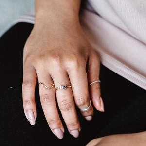 May include: A hand wearing three gold rings with small gemstones. The rings are on the middle and ring fingers. The hand is resting on a black surface.