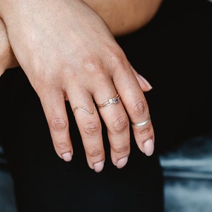May include: A hand wearing three gold rings. The ring on the middle finger has a small blue gemstone. The ring on the index finger is a thin, wavy band. The ring on the pinky finger is a simple, thin band.