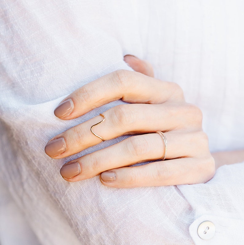 May include: A hand wearing two gold rings. The ring on the index finger is a thin, wavy band. The ring on the middle finger is a simple, thin band.