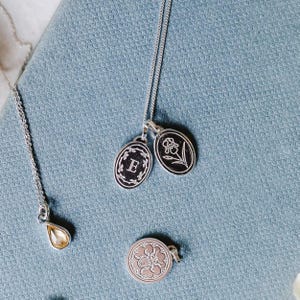 May include: Silver necklaces with pendants. One pendant is a teardrop shape with a yellow gemstone. Two oval pendants are on a chain, one with the letter "E" and the other with a floral design. A round pendant with a floral design is also visible.