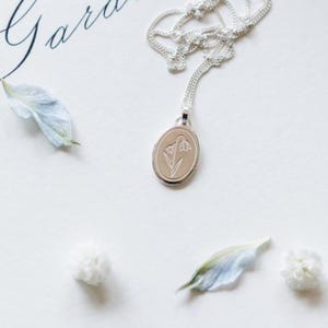 May include: A silver necklace with a delicate oval pendant featuring an engraved snowdrop flower design. The pendant hangs on a silver chain.