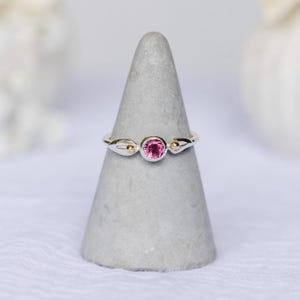 May include: A delicate ring with a silver band and a vibrant pink gemstone at the center. The ring features small gold accents on either side of the central stone, adding a touch of elegance. The ring is displayed on a gray, cone-shaped stand.