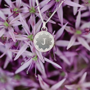 May include: A silver pendant necklace with a floral design and the letter "J" in the center. The pendant hangs on a delicate chain, set against a backdrop of purple flowers. The necklace is a piece of jewelry.