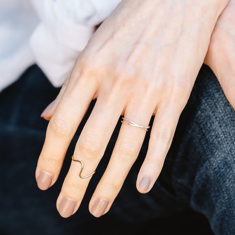 May include: A close-up of a hand wearing two thin gold rings. The ring on the index finger is a simple band, while the ring on the middle finger is a wavy design.