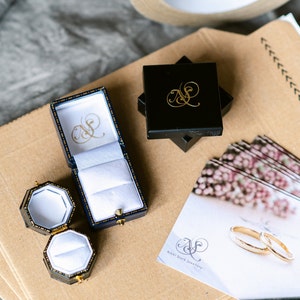 May include: Two black ring boxes with gold lettering, one open with a white interior, and one closed. A smaller black ring box with a white interior is open. A white card with gold lettering that reads "Nikki Stark Jewellery" and a floral design is on a brown surface.