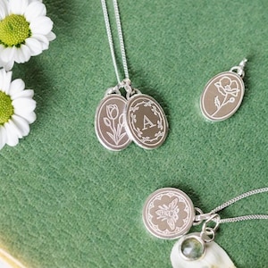 May include: A silver necklace with three oval charms. The charms feature a tulip, a letter A, and a poppy. The necklace is on a green surface.