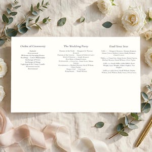 May include: A white rectangular wedding program card featuring black printed text titled Order of Ceremony, The Wedding Party, and Find Your Seat, surrounded by white roses, green eucalyptus sprigs, a gold pen, and a light pink silk ribbon.