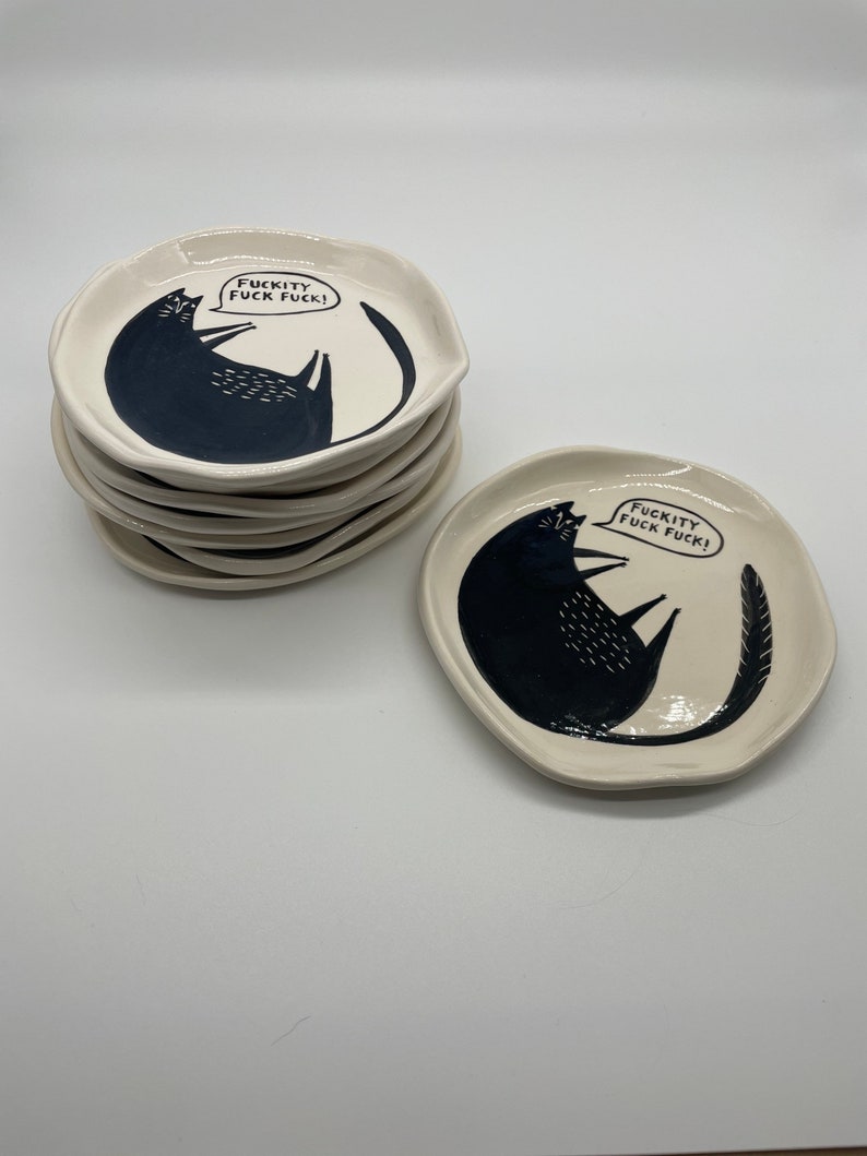 Może przedstawiać: Jasne ceramiczne talerze z czarną ilustracją kota. Każdy talerz ma czarny wz&oacute;r kota i tekst "FUCKITY FUCK FUCK!" w dymku. Talerze mają falisty brzeg. Widoczny jest stos talerzy.