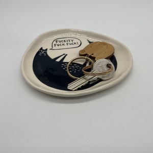 Może przedstawiać: Mały, kremowy ceramiczny talerz z czarną ilustracją kota i napisem "FUCKITY FUCK FUCK!". Na talerzu znajdują się klucze i drewniany brelok. Kot leży z podniesionymi ramionami. Talerz ma lekko nieregularny kształt.