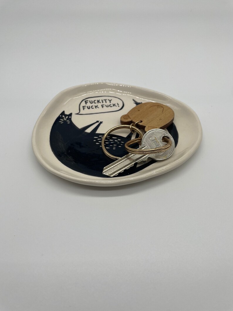 Może przedstawiać: Mały, kremowy ceramiczny talerz z czarną ilustracją kota i napisem "FUCKITY FUCK FUCK!". Na talerzu znajdują się klucze i drewniany brelok. Kot leży z podniesionymi ramionami. Talerz ma lekko nieregularny kształt.