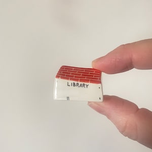 May include: A miniature ceramic library figurine. The roof is red brick, and the front is white with the word "LIBRARY" in black lettering. The figurine is held between two fingers against a white background.
