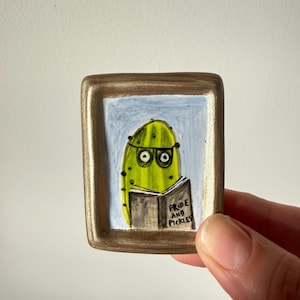 Könnte beinhalten: Eine kleine rechteckige Keramikplakette mit einem metallisch goldenen Rahmen, die eine handgemalte Illustration einer grünen Gewürzgurke mit Brille zeigt, die ein Buch mit dem Titel Pride and Pickles vor hellblauem Hintergrund liest.