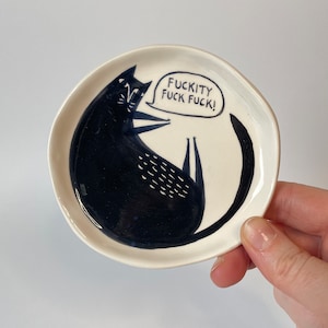 Może przedstawiać: Mały, okrągły, kremowy ceramiczny talerz z ilustracją czarnego kota. Kot leży z podwiniętym ogonem. Dymek zawiera tekst "FUCKITY FUCK FUCK!". Talerz jest trzymany przez dłoń.