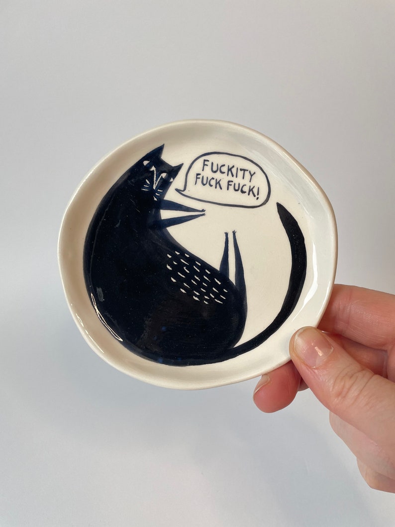 Może przedstawiać: Mały, okrągły, kremowy ceramiczny talerz z ilustracją czarnego kota. Kot leży z podwiniętym ogonem. Dymek zawiera tekst "FUCKITY FUCK FUCK!". Talerz jest trzymany przez dłoń.