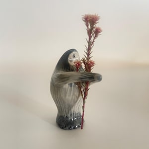 May include: A small, hand-painted ceramic figurine of a grey and white creature with a rounded body and dark base. The figure has a simple face with two dots for eyes and holds a tall, slender stem with small, pinkish-red flower clusters.
