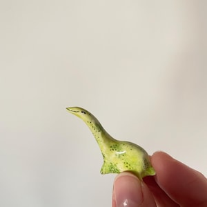 May include: A small, green ceramic figurine of a plesiosaur with a long neck and a spotted pattern.