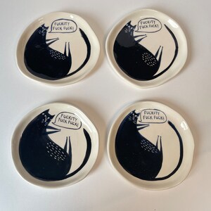Może przedstawiać: Cztery kremowobiałe ceramiczne talerze z ilustracją czarnego kota. Każdy talerz przedstawia czarnego kota z tekstem "FUCKITY FUCK FUCK!" w dymku. Talerze mają lekko nieregularny, organiczny kształt.