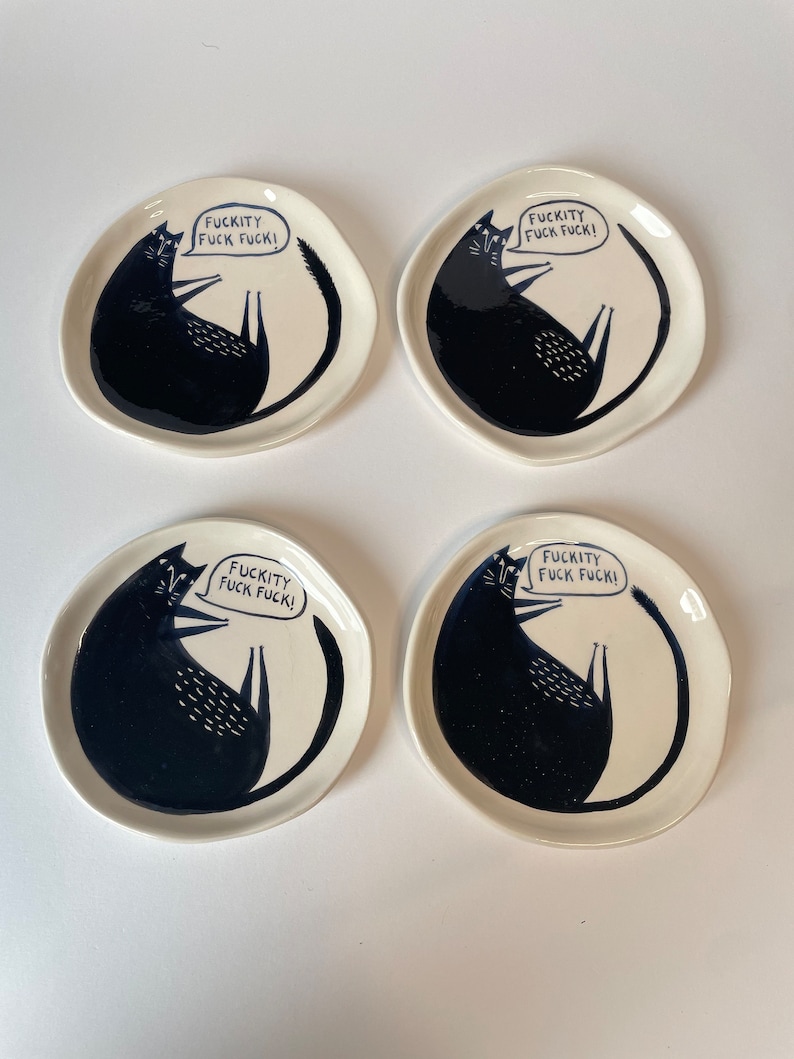 Może przedstawiać: Cztery kremowobiałe ceramiczne talerze z ilustracją czarnego kota. Każdy talerz przedstawia czarnego kota z tekstem "FUCKITY FUCK FUCK!" w dymku. Talerze mają lekko nieregularny, organiczny kształt.