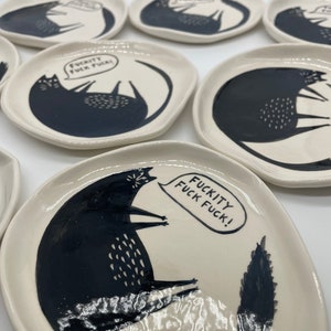 Może przedstawiać: Zestaw małych, kremowych ceramicznych talerzy. Każdy talerz ma czarną ilustrację kota z tekstem "FUCKITY FUCK FUCK!" w dymku. Kot leży z podwiniętym ogonem.