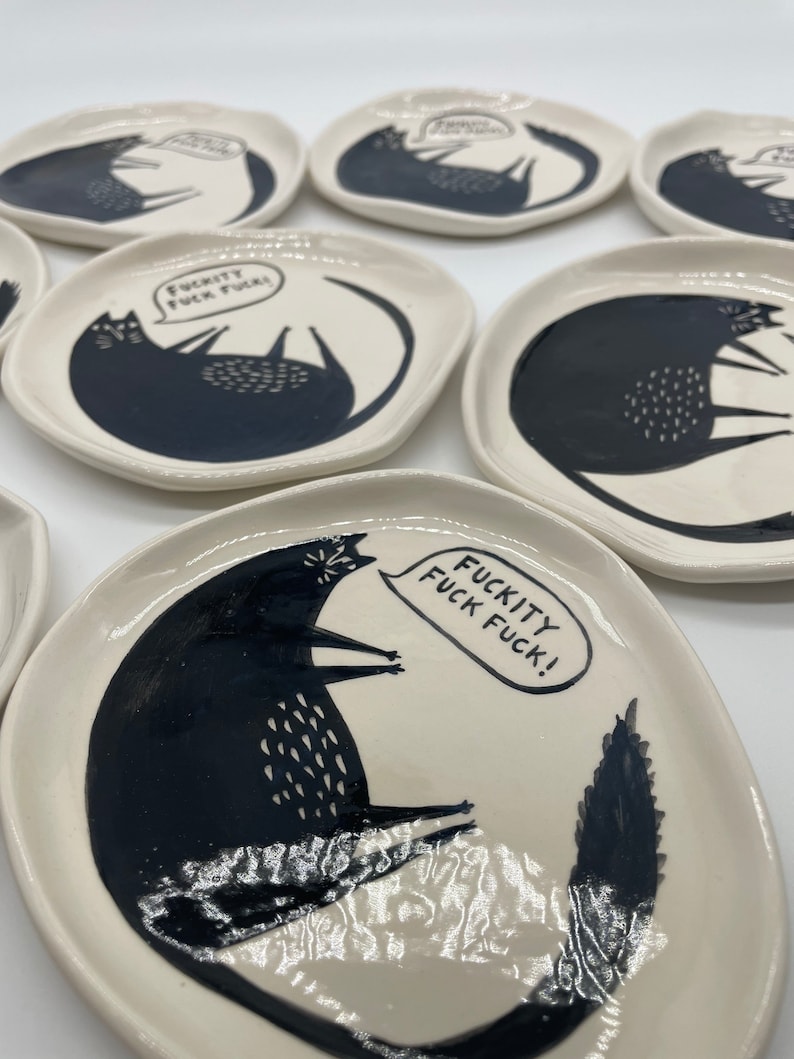 Może przedstawiać: Zestaw małych, kremowych ceramicznych talerzy. Każdy talerz ma czarną ilustrację kota z tekstem "FUCKITY FUCK FUCK!" w dymku. Kot leży z podwiniętym ogonem.