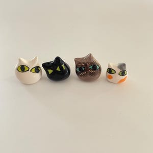 May include: Four ceramic cat head figurines in white, black, brown, and white with orange spots. Each cat has detailed eyes in various colours, including yellow, green, and blue. The figurines are small and likely decorative.