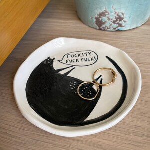 Może przedstawiać: Biała ceramiczna patera na drobiazgi z czarną ilustracją kota i napisem "FUCKITY FUCK FUCK!". Na paterze leżą dwa złote pierścionki. W tle znajduje się jasnoniebieski ceramiczny garnek.