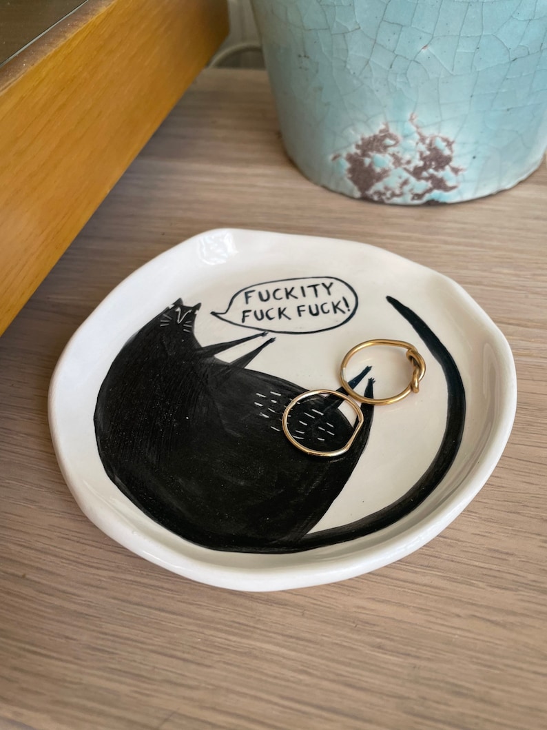 Może przedstawiać: Biała ceramiczna patera na drobiazgi z czarną ilustracją kota i napisem "FUCKITY FUCK FUCK!". Na paterze leżą dwa złote pierścionki. W tle znajduje się jasnoniebieski ceramiczny garnek.