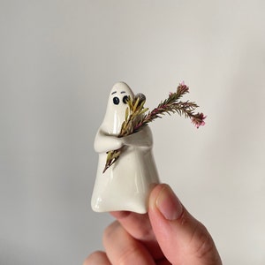 Könnte beinhalten: Weiße Keramik-Geisterfigur, die einen Strauß getrockneter rosa Blumen hält. Der Geist hat schwarze Augen und ein einfaches Lächeln.