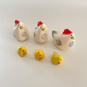 Puede incluir: Un conjunto de cinco figuritas de cerámica de pollos. Tres son blancas con crestas rojas y marcas negras. Dos son amarillas con picos naranjas y ojos negros.